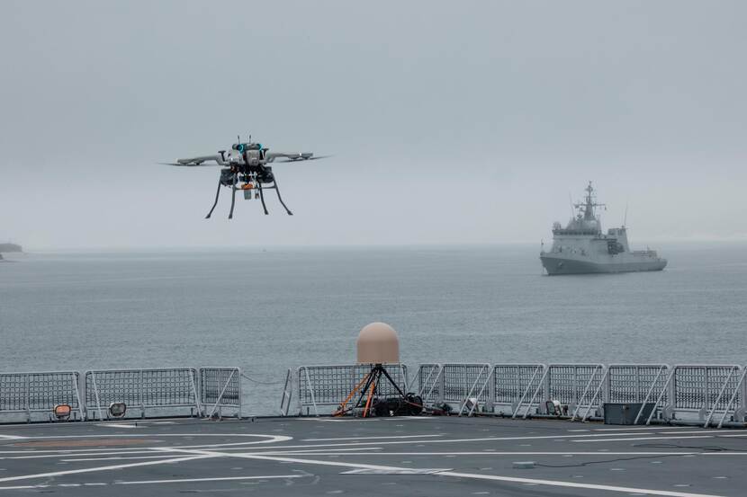 Een drone vliegt boven het dek van een marineschip. Op de achtergrond een marineschip op zee.