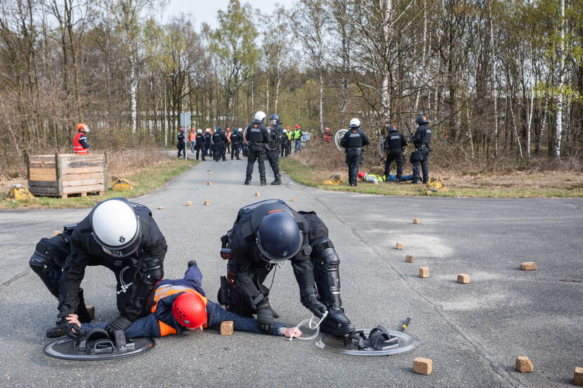2 leden van de BE drukken een persoon tegen de grond. Op de achtergrond houden andere marechaussees een groep mensen op afstand.