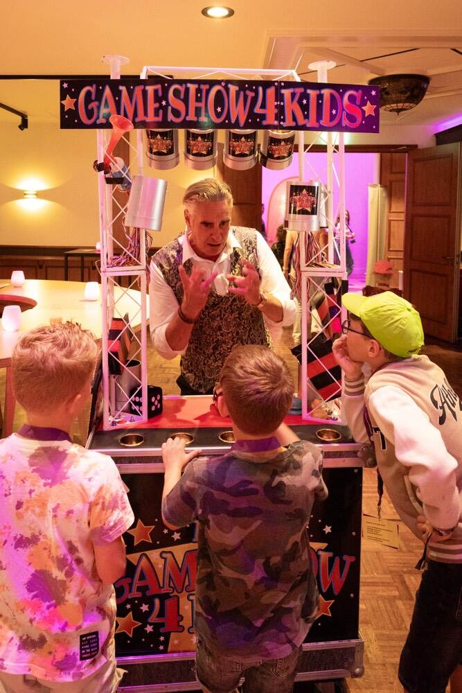 Een man achter een stand met de tekst: Gameshow 4 kids. Drie kinderen kijken toe.